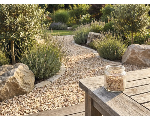 Gartenweg aus beigem Kies, gesäumt von Lavendel, Olivenbäumen und Findlingen, mit einem Glas Kies auf einem Holztisch.