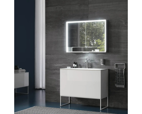 Modernes Badezimmer mit weißem Waschtischunterschrank auf Metallgestell, Waschbecken mit Chromarmatur und LED-Spiegelschrank vor dunklen Wänden.