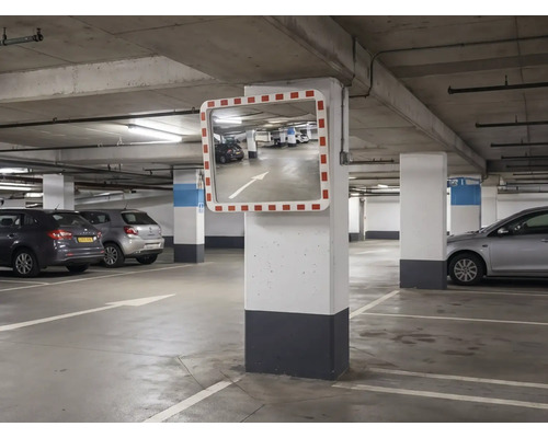 Rechteckiger Verkehrsspiegel mit rot-weißem Rahmen, montiert an einer Betonsäule in einer hellen, organisierten Tiefgarage mit geparkten Autos.