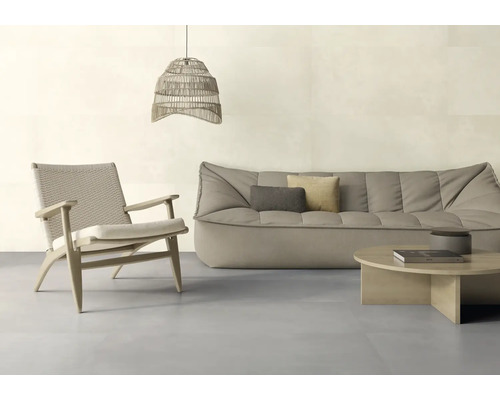Ein helles, modern eingerichtetes Wohnzimmer mit einem niedrigen Stoffsofa, einem gewebten Sessel, einem runden Holztisch und einer Pendelleuchte.