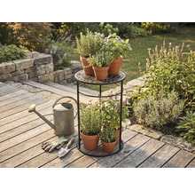 Zweistöckiges Pflanzenregal aus Metall mit Kräutern in Terrakotta-Töpfen auf einer Holzterrasse neben einer Gießkanne und Gartenwerkzeug.
