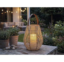 Laterne aus Korbgeflecht mit Henkel und brennender Kerze auf einem Holztisch in einem beleuchteten Garten mit Lounge-Möbeln.