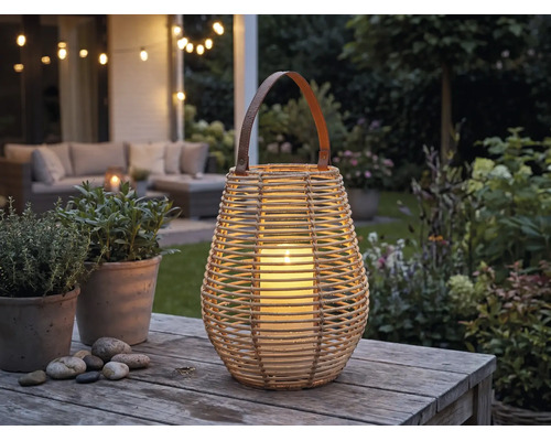 Laterne aus Korbgeflecht mit Henkel und brennender Kerze auf einem Holztisch in einem beleuchteten Garten mit Lounge-Möbeln.