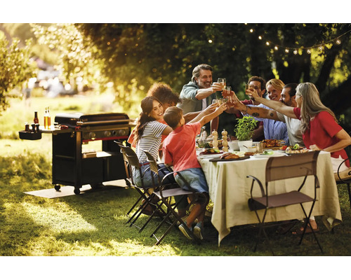 Eine Gruppe von Menschen bei einer Gartenparty mit einem Gasgrill im Hintergrund, die an einem gedeckten Tisch im Freien anstoßen.