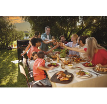Gruppe von Personen beim Essen an einem langen Tisch im Garten mit Gasgrill und Lichterkette im Hintergrund.