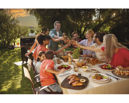 Gruppe von Personen beim Essen an einem langen Tisch im Garten mit Gasgrill und Lichterkette im Hintergrund.
