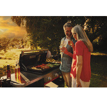 Ein Paar grillt in einem hellen Garten bei Sonnenuntergang auf einer modernen Gas-Grillstation mit Fleisch und Gemüse.