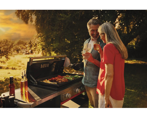 Ein Paar grillt in einem hellen Garten bei Sonnenuntergang auf einer modernen Gas-Grillstation mit Fleisch und Gemüse.