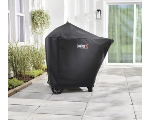 Schwarze Abdeckhaube für einen Grill mit Weber Logo auf einer hellen Steinterrasse im Außenbereich neben Pflanzen.