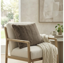 Quadratisches, geripptes Dekokissen in Felloptik auf einem hellen Sessel mit Strickdecke in einem hellen, modern eingerichteten Raum.