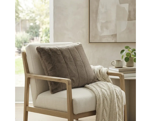 Quadratisches, geripptes Dekokissen in Felloptik auf einem hellen Sessel mit Strickdecke in einem hellen, modern eingerichteten Raum.