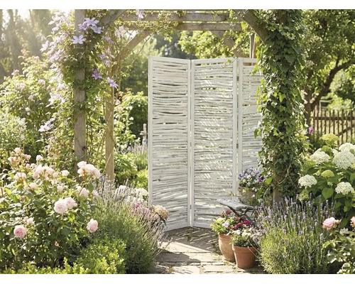 Dreiteiliger weißer Paravent aus Holzästen in einem hellen Garten mit Rosen, Lavendel und einem mit Rankpflanzen bewachsenen Torbogen.