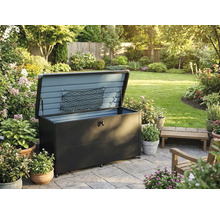 Garten-Auflagenbox aus Metall mit geöffnetem Deckel und Aufbewahrungsnetz auf einer gepflasterten Terrasse in einem Garten mit Pflanzen und Blumen.