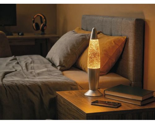 Glitzer-Lavalampe aus Metall auf einem hölzernen Nachttisch in einem Schlafzimmer neben einem Bett.