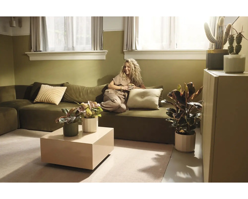 Helles Wohnzimmer mit olivgrünem Sofa, Frau mit Tasse, beigefarbenem Couchtisch und verschiedenen Zimmerpflanzen in natürlichem Stil.