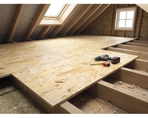Dachbodenausbau mit OSB-Platten auf Holzbalkenlage, Akkuschrauber und Maßband auf der Arbeitsfläche im hellen Dachstock.