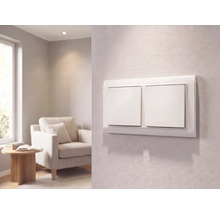 Moderner weißer Zweifach-Lichtschalter an einer strukturierten Wand in einem hellen Wohnzimmer mit Sessel und Beistelltisch.