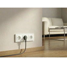 Dreifach-Steckdose mit eingestecktem Stecker an einer Wand über Holzboden in einem hellen Wohnzimmer.