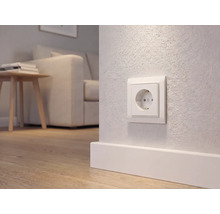 Weiße Schutzkontakt-Steckdose an einer hellen Wand über einer Fußleiste in einem modern eingerichteten Wohnzimmer mit Holzboden.