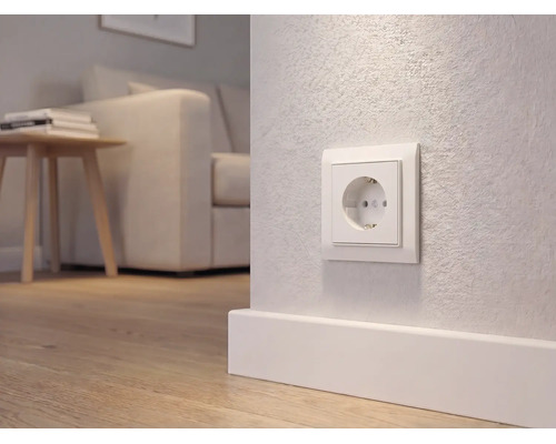 Weiße Schutzkontakt-Steckdose an einer hellen Wand über einer Fußleiste in einem modern eingerichteten Wohnzimmer mit Holzboden.