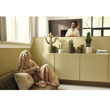 Modernes, helles Wohnzimmer mit Kakteen in Pflanztöpfen auf einem Sideboard, einer Frau auf dem Sofa und einem Mann im Hintergrund.