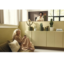Helles Wohnzimmer mit Frau auf Sofa und Mann hinter Trennwand. Kakteen in Töpfen stehen auf einem minimalistischen Schrank.