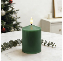 Grüne LED-Stumpenkerze mit realistischer Flamme auf weißem Marmortisch mit Eukalyptus. Hell gestalteter Raum mit einem Weihnachtsbaum.