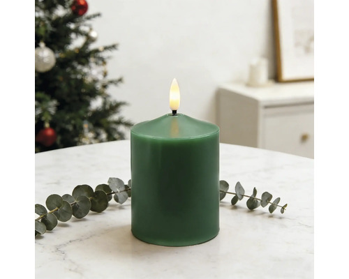 Grüne LED-Stumpenkerze mit realistischer Flamme auf weißem Marmortisch mit Eukalyptus. Hell gestalteter Raum mit einem Weihnachtsbaum.