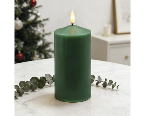 Dunkelgrüne LED-Stumpenkerze mit flackernder Flamme auf hellem Tisch mit Eukalyptuszweig in einem hellen, weihnachtlichen Raum.
