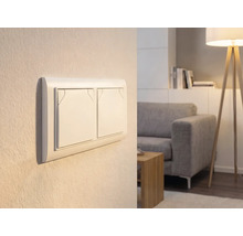 Doppel-Lichtschalter an einer Wand in einem hellen Wohnzimmer mit Sofa und Stehlampe.