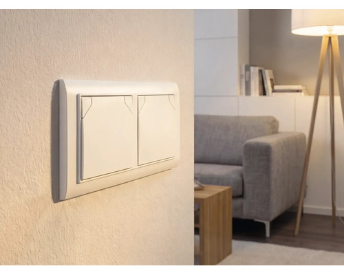 Doppel-Lichtschalter an einer Wand in einem hellen Wohnzimmer mit Sofa und Stehlampe.