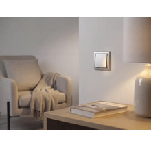 Moderner silberner Wippschalter an der Wand in einem hellen Wohnzimmer mit Sessel und Tischlampe.