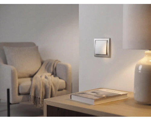 Moderner silberner Wippschalter an der Wand in einem hellen Wohnzimmer mit Sessel und Tischlampe.