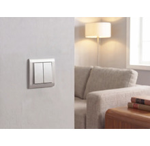 Moderner Doppel-Lichtschalter an einer Wand in einem hellen Wohnzimmer mit Sofa, Stehlampe und Holztisch.