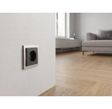 Moderne Steckdose mit silbernem Rahmen und schwarzem Einsatz in einem hellen Wohnzimmer mit Parkettboden und Sofa.