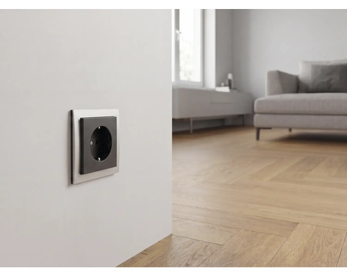 Moderne Steckdose mit silbernem Rahmen und schwarzem Einsatz in einem hellen Wohnzimmer mit Parkettboden und Sofa.