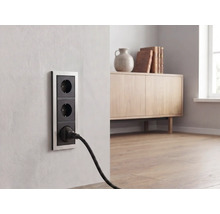 Dreifache Wandsteckdose mit Metallrahmen in einem hellen Wohnzimmer, ein schwarzer Stecker ist eingesteckt.