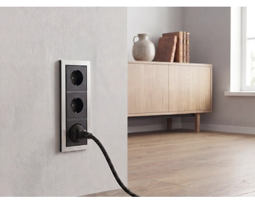 Dreifache Wandsteckdose mit Metallrahmen in einem hellen Wohnzimmer, ein schwarzer Stecker ist eingesteckt.