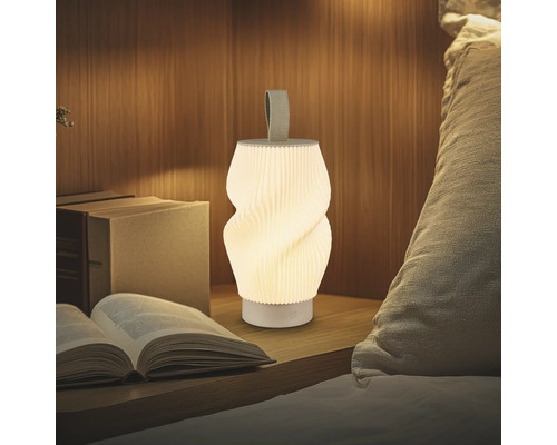 Tragbare LED-Leuchte mit Plissee-Design und Textilschlaufe auf einem Nachttisch aus Holz neben Büchern und Kissen. Warm beleuchtet.