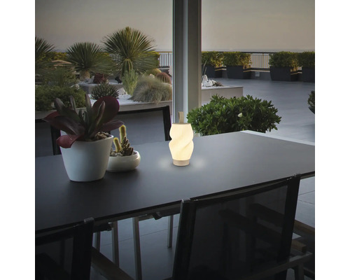 Moderne weiße LED-Tischleuchte im Spiraldesign auf einem dunklen Terrassentisch, umgeben von Topfpflanzen bei Dämmerung.