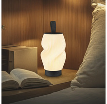 Moderne tragbare LED-Tischleuchte mit gedrehtem geripptem Schirm und schwarzem Griff auf einem Nachttisch neben Büchern und einem Kissen.