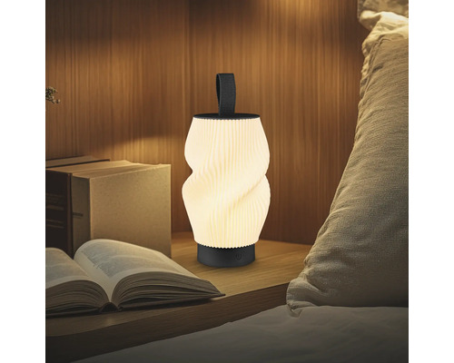 Moderne tragbare LED-Tischleuchte mit gedrehtem geripptem Schirm und schwarzem Griff auf einem Nachttisch neben Büchern und einem Kissen.