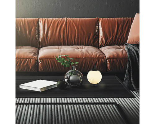 Moderne, kugelförmige Tischleuchte auf einem schwarzen Couchtisch vor einem braunen Ledersofa in einem stilvoll eingerichteten Wohnzimmer.