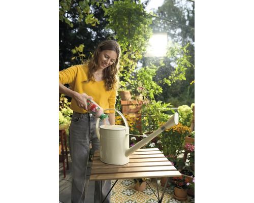 Eine Frau füllt Flüssigdünger aus einer Flasche in eine Metallgießkanne auf einem Holztisch in einem hellen Garten mit Pflanzen.