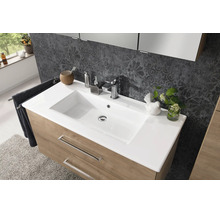 Moderner Waschtisch mit weißem Waschbecken, verchromter Armatur und Unterschrank in Holzoptik vor einer gemusterten Wand.
