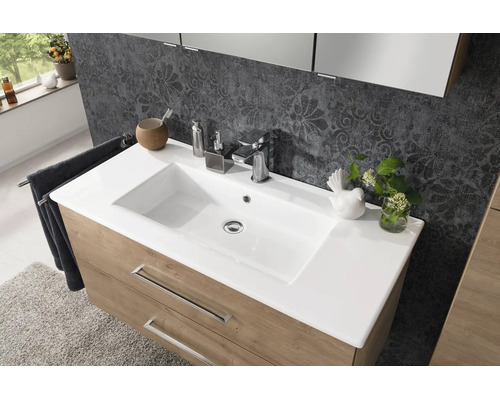 Moderner Waschtisch mit weißem Waschbecken, verchromter Armatur und Unterschrank in Holzoptik vor einer gemusterten Wand.