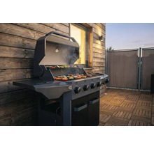 Moderner Gasgrill mit geöffnetem Deckel auf einer Holzterrasse, Grillgut auf dem Rost vor einer Holzhauswand.