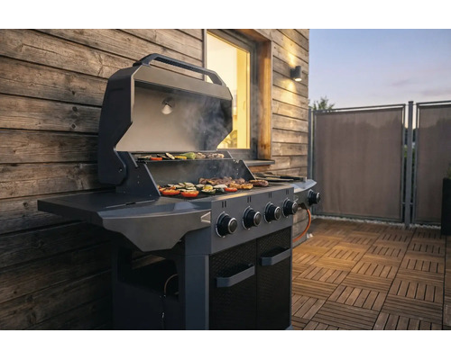 Moderner Gasgrill mit geöffnetem Deckel auf einer Holzterrasse, Grillgut auf dem Rost vor einer Holzhauswand.