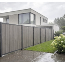 Moderner Sichtschutzzaun aus vertikalen Verbundwerkstoff-Paneelen bei Regen in einem Garten mit Rasen vor einem Wohnhaus.