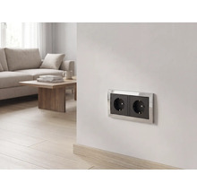 Zweifache Schutzkontakt-Steckdose mit schwarzen Einsätzen und silbernem Rahmen an einer Wand in einem hellen Wohnzimmer.